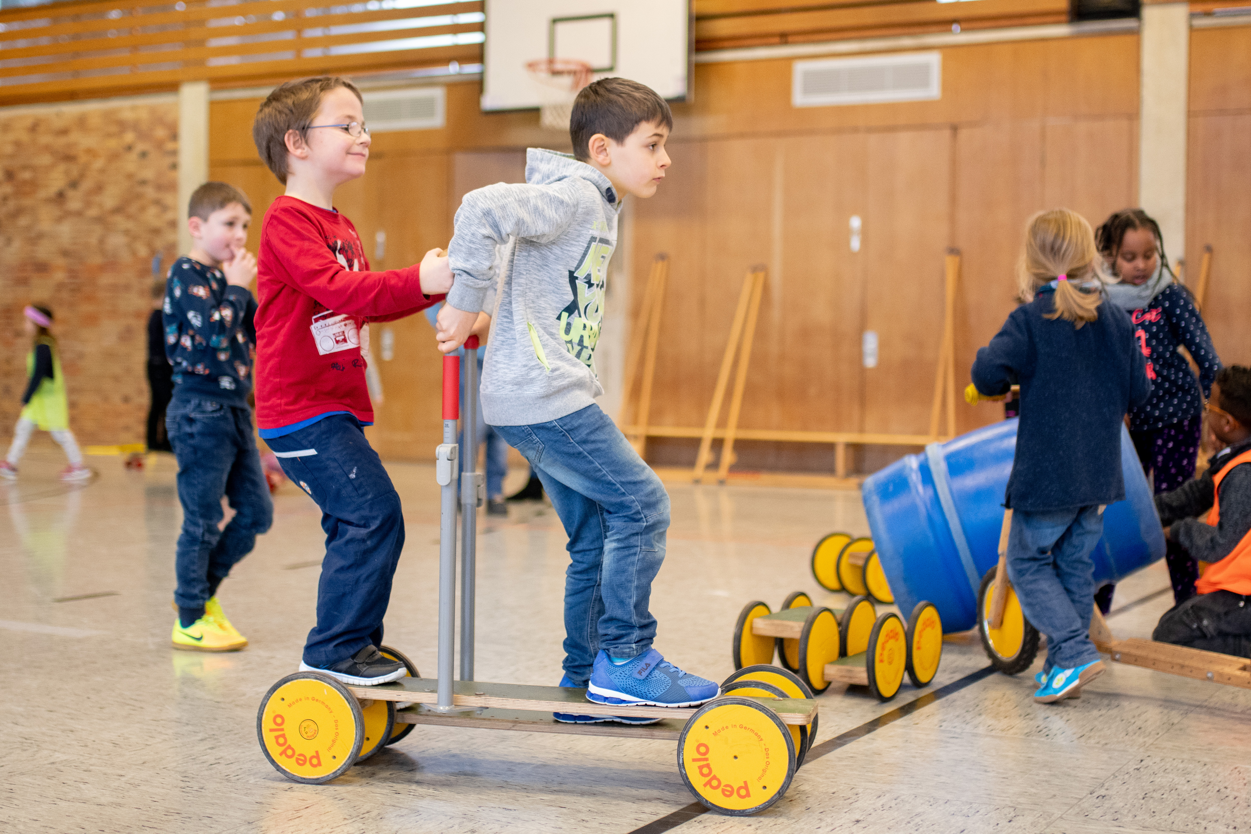 Zwei Jungs stehen auf einer Art Roller und fahren zusammen durch die Halle.