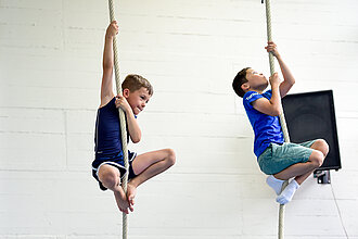 [Translate to Einfache Sprache:] Zwei Jungs klettern in einer Sporthalle Seile hinauf.