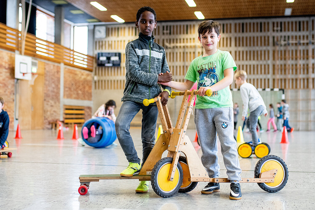 Zwei Kinder mit Tretroller in der Turnhalle.