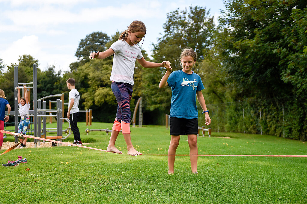 Kind balanciert auf einer Slack-Line im Garten, ein anderes Kind hält die Hand.