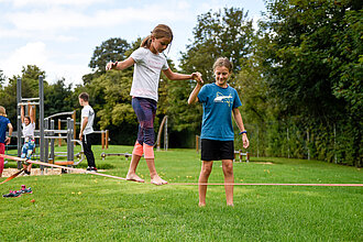 Kind balanciert auf einer Slack-Line im Garten, ein anderes Kind hält seine Hand.