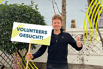 Mann hält ein Schild hoch mit Aufschrift: Volunteers gesucht. 