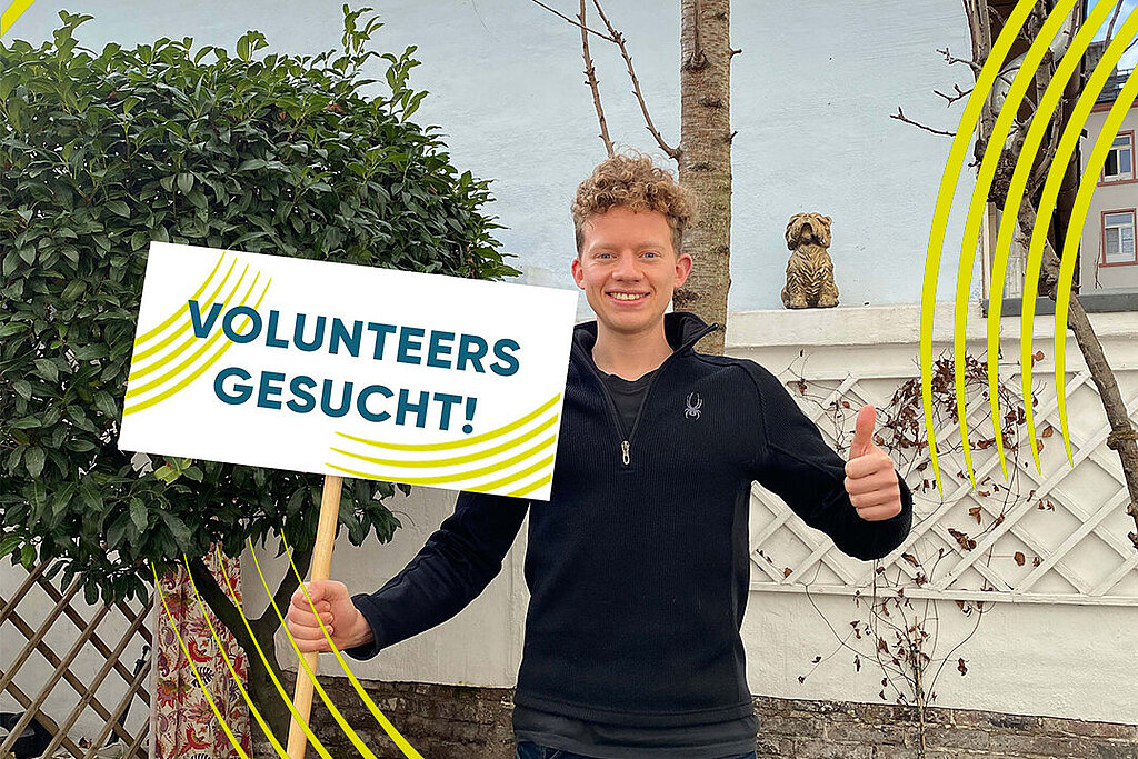 [Translate to Einfache Sprache:] Mann hält ein Schild hoch mit Aufschrift: Volunteers gesucht. 