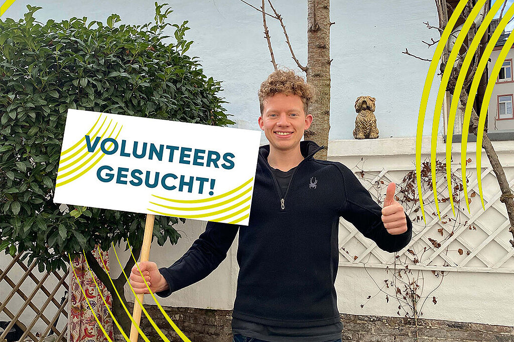 Mann hält ein Schild hoch mit Aufschrift: Volunteers gesucht. 