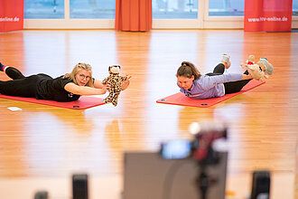 Zwei Frauen machen eine Sportübung, eine Frau hält ein Kuscheltier.