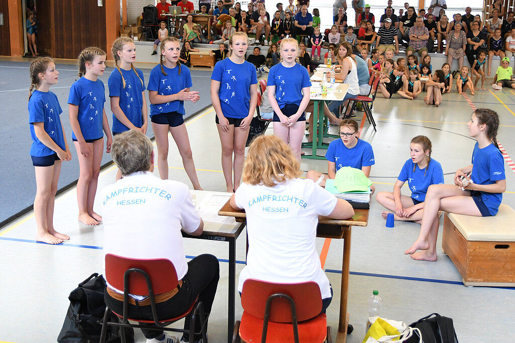 [Translate to Einfache Sprache:] Eine Kindergruppe singt vor zwei Kampfrichterinnen.