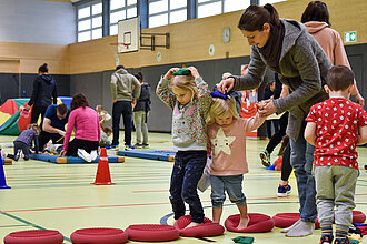 Kinder balancieren in einer Turnhalle gemeinsam mit einer Frau über Balance-Kissen.