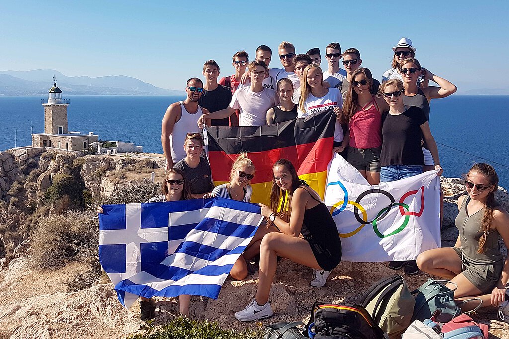 Mehrere Jugendliche halten eine deutsche, eine griechische und eine olympische Flagge und lachen in die Kamera.