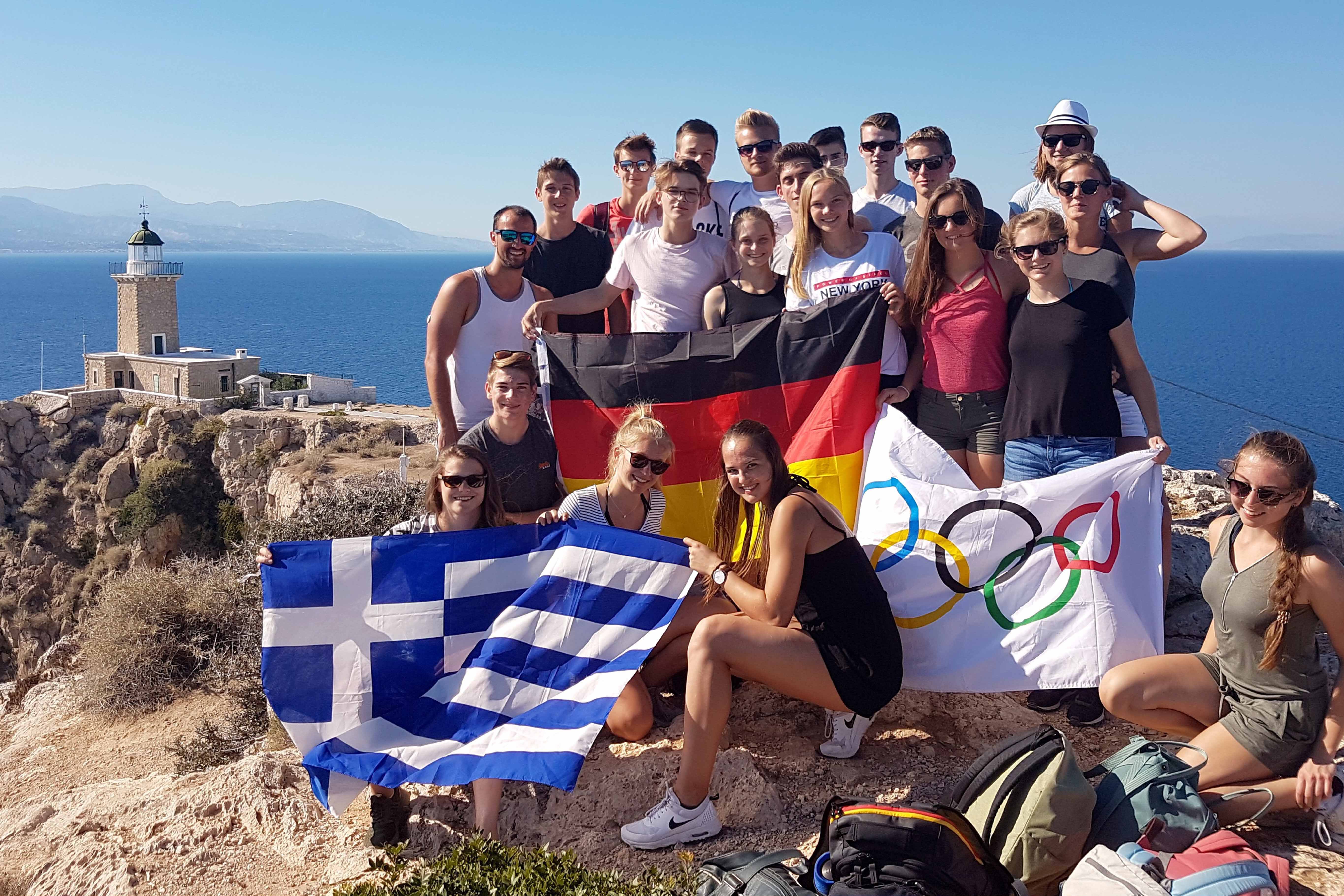 Mehrere Jugendliche halten eine deutsche, eine griechische und eine olympische Flagge und lachen in die Kamera.