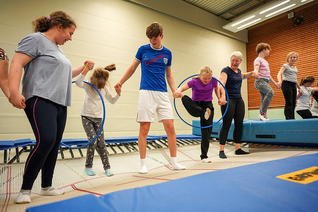 Eine Gruppe von Menschen steht auf dem Trampolin und steigen einzeln durch einen Gymnastikreifen.