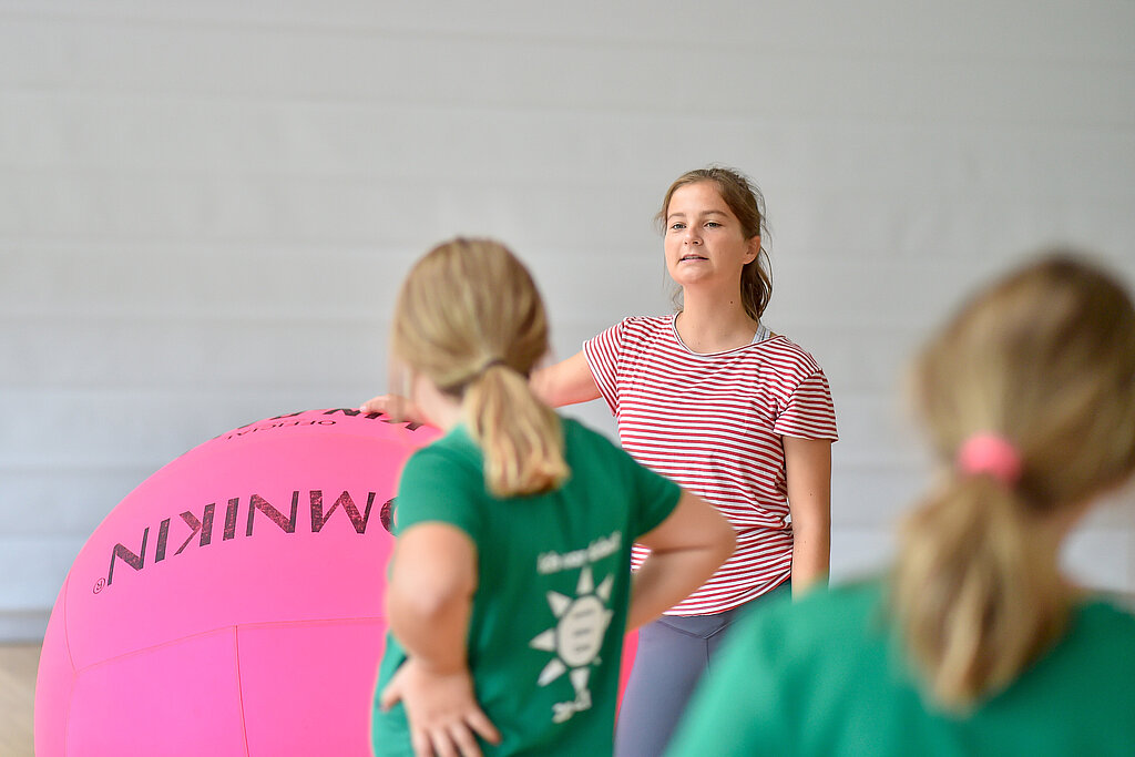 [Translate to Einfache Sprache:] Junge Frau steht in der Halle und spricht zu Kindern. 