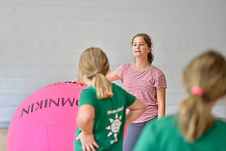 [Translate to Einfache Sprache:] Junge Frau steht in der Halle und spricht zu Kindern. 