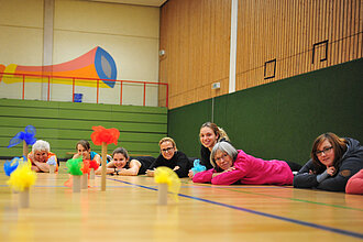 Frauen liegen auf dem Hallenboden und spielen ein Spiel mit bunten Tüchern.