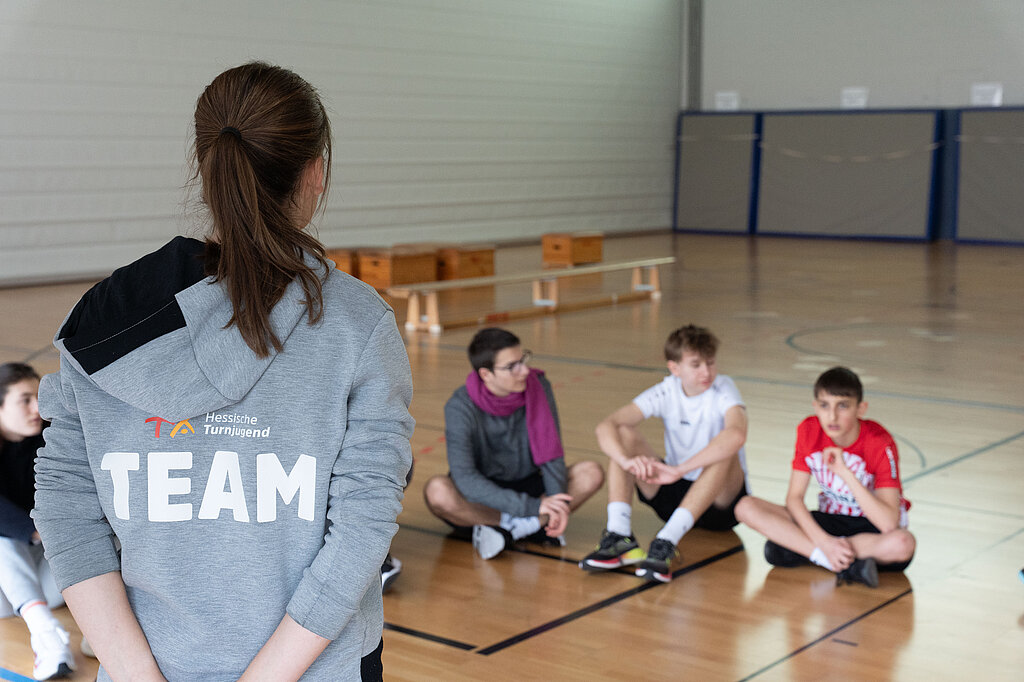Eine Junge Frau mit Jacke erklärt Schülern etwas in einer Sporthalle.