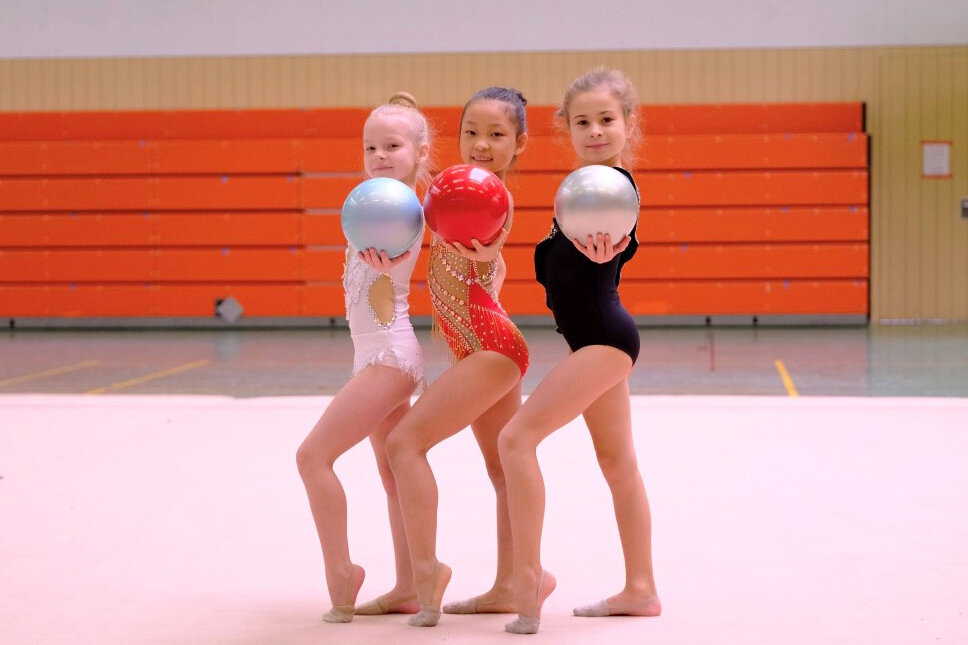 Drei Mädchen der Rhythmischen Sportgymnastik halten einen Gymnastikball in die Kamera.