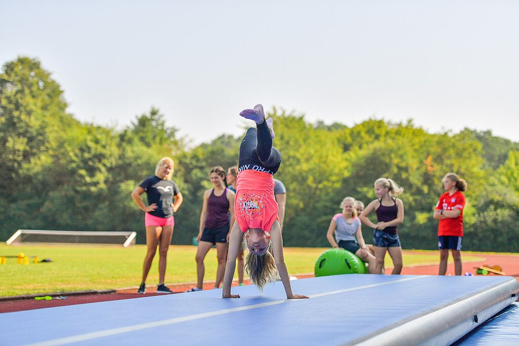 Ein Mädchen turnt draußen und mehrere Kinder schauen zu.
