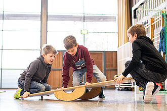 Drei Jungs spielen in einer Turnhalle auf einer Wippe.