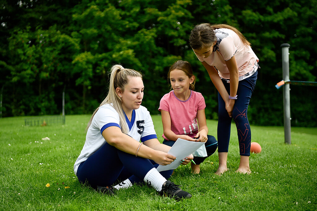 Junge Frau sitzt mit zwei Mädchen im Garten und bespricht eine Aufgabe.