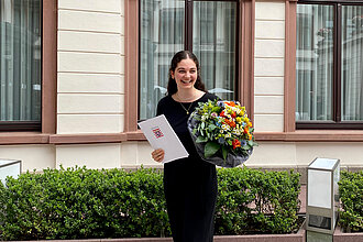 Eine Frau hat einen Blumenstrauß und eine Ehrenurkunde in der Hand und lacht in die Kamera.