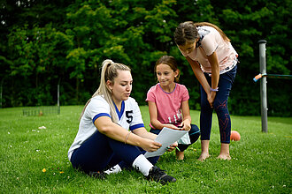 Eine Frau sitzt auf einer grünen Wiese und erklärt zwei jungen Mädchen einen Zettel.
