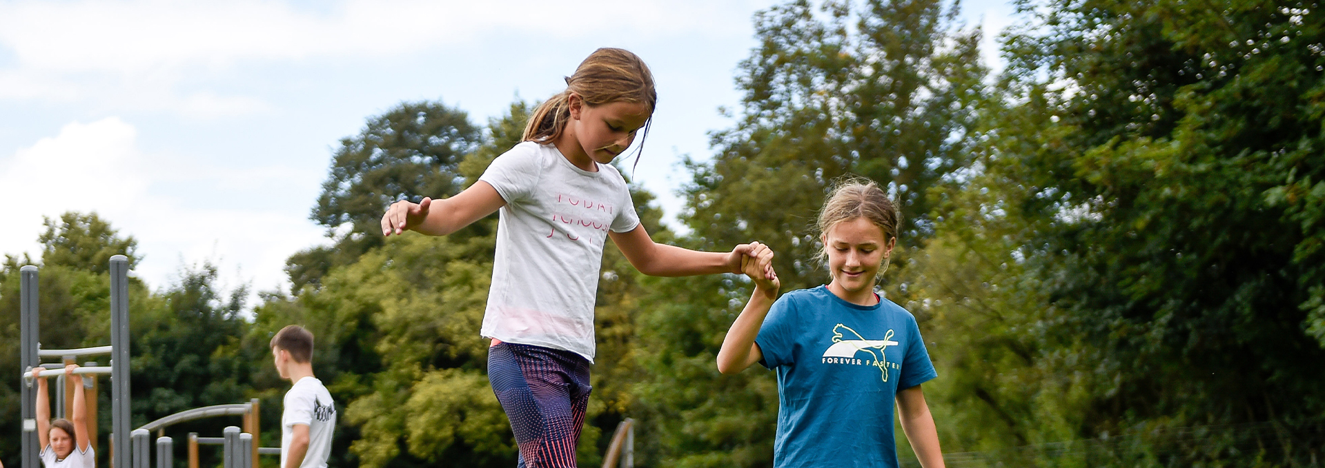 Kind balanciert auf einer Slackline und wird von einem anderen Kind an der Hand gehalten.