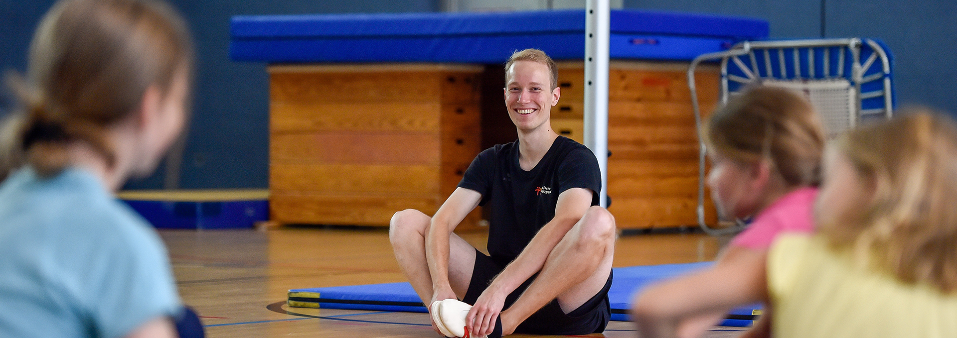 Ein Übungsleiter sitzt mit Kindern im Kreis in einer Sporthalle.