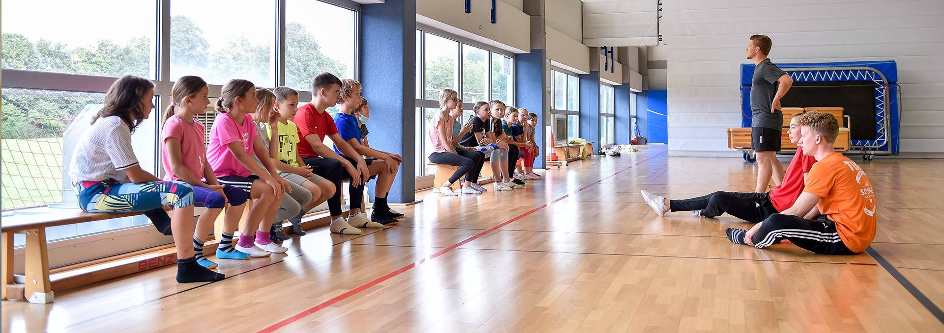 Übungsleiter sitzen Kindern auf einer Bank in einer Sporthalle gegenüber.